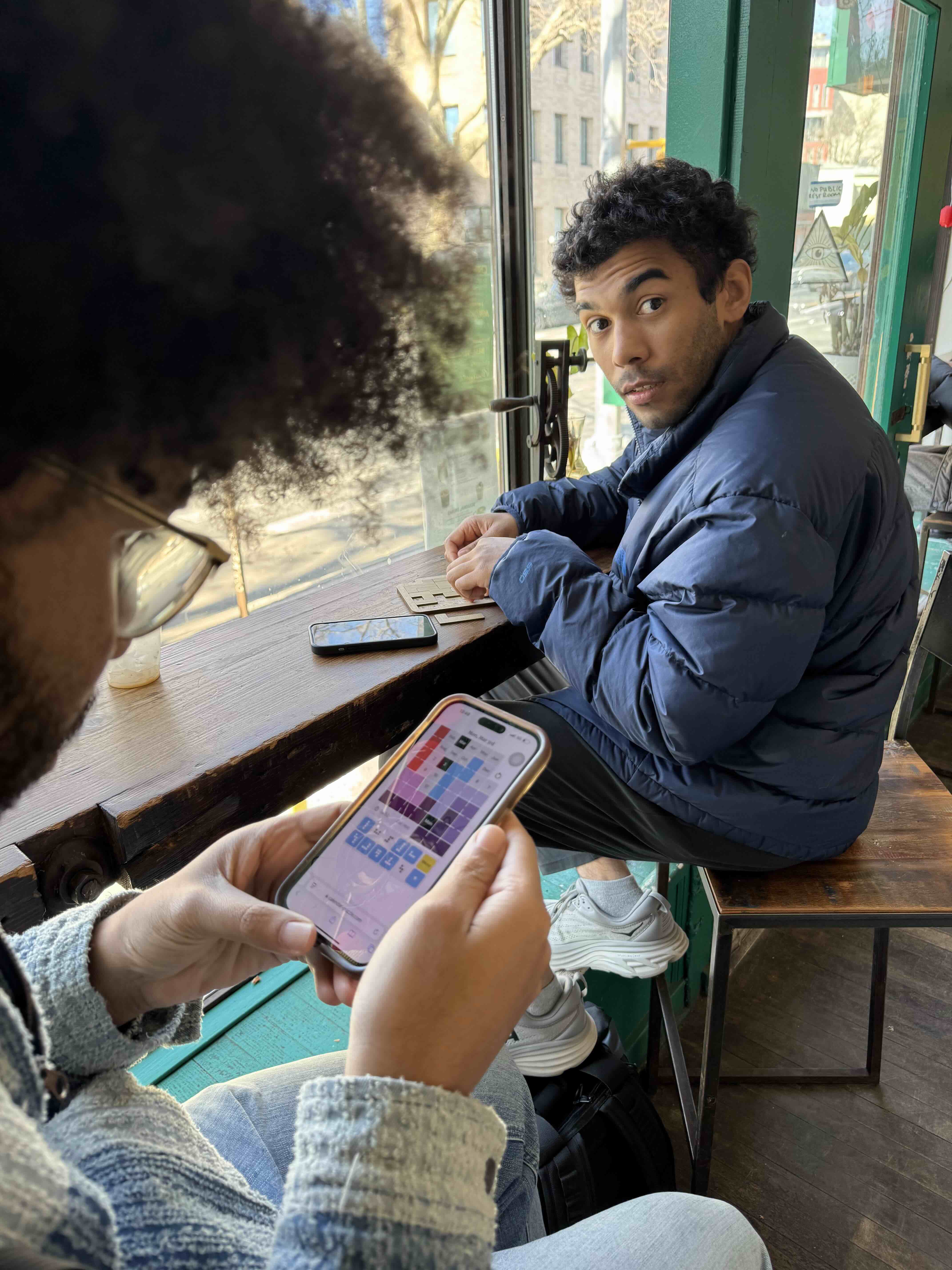 photo of two people in a coffee shop, one playing the digital calendar puzzle game and one playing the physical game.